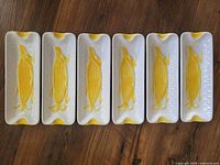 Photo of all six ceramic corn on the cob dishes, showing the top view and arrangement in a row. Each dish is white with a yellow raised corn cob design and yellow corner accents.