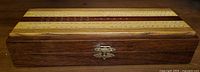 Closed wooden cribbage board box showing top side with pegboard holes in alternating wood stripes, secured with brass clasp.