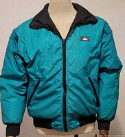 Front view of Woolrich teal reversible bomber jacket displayed on mannequin, showing zip front, rib-knit cuffs and waistband, and small brand patch on chest.