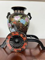 Front and angled view of black ceramic vase with colorful peacock and floral designs on a decorative stand with two black and red ornamental wheels.