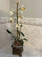 Full view of the artificial orchid plant standing approximately 2-3 feet high, with five open orchid blooms and two unopened buds on a single green stem. The square pot has ornate classical designs and is likely made of resin or ceramic.