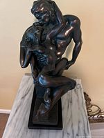 Front view of dark bronze resin sculpture showing a nude male seated and holding a nude female in an intimate embrace on a black base.