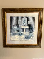 Framed square print artwork showing an interior scene with furniture in blue tones mounted in a wood gold-finish frame.