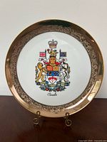 Full view of the porcelain plate showing the Canadian coat of arms at the center, gold rim, and ornate edge detailing on a display stand.