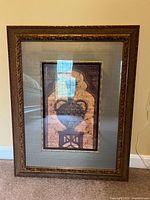 Framed art print featuring a classical urn or vase with an ornate wood frame and wide fabric mat, shown hanging on a wall with visible window reflections on glass.