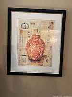 Framed wall art showing Asian-style vase with calligraphy and decorative stamps on beige background, framed in black wood or composite with glass.