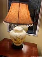 Full view of the oriental ceramic table lamp on a wooden surface, showing ceramic base with nature motif and fabric lampshade with fringe.
