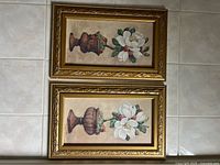 Full view of the pair of framed floral paintings showing vases with white flowers in ornate gold-tone frames