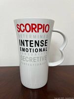 Front side of white ceramic mug with red 'SCORPIO' text and zodiac trait descriptions in black and gray. Heart-shaped handle visible in profile.