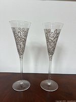 Two tall champagne flutes with floral etched frosted design on the upper part against white background on wood surface.