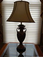 Full view of the table lamp including shade and base on glass surface in front of window blinds.