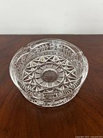 Clear pressed glass ashtray with starburst and floral design, resting on wooden surface.