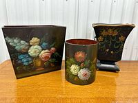 Photo showing three wooden waste paper baskets or planters painted with floral designs arranged on a table against a white wall.