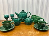 Overview of the entire pottery tea set on wooden surface including teapot, sugar bowl, creamer, cups and saucers, goblets, and mugs.