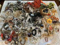 Overview photo of mixed assortment of costume jewelry laid out on a white patterned cloth showing many necklaces, bracelets, earrings, and rings in various styles and materials.