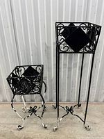 Pair of black wrought iron garden planters standing side by side on concrete floor against corrugated metal background, showcasing size difference and paint wear.