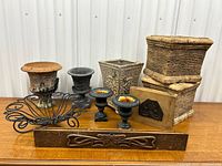 Front view of entire lot showing all items on wooden table under white wall: three cast iron urns, two plaster planters, one ornate gray square planter, metal bowl, wood plaque and antique pine drawer.