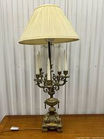 Full view of the tall vintage ornate lamp with cream fabric shade, gilt brass candelabra arms, and detailed lion head urn base on a wood surface.