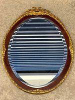Full view of oval wall mirror featuring walnut frame with gold gilt decorative trim and clear beveled glass reflecting horizontal blinds and surroundings.
