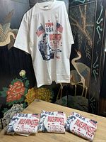 One shirt displayed on hanger showing front graphic; three folded identical shirts below