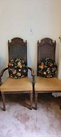 Two vintage dining chairs with dark wood frames, cane backs, gold seats, and two brown floral pillows placed on them, positioned side by side on carpet.