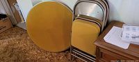Round yellow foam topped card table leaning against the wall beside the 4 stacked folding chairs