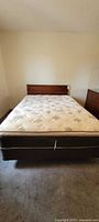 Front view of full-size mattress atop box spring and wood headboard visible against beige wall with carpeted floor.