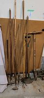 Various long-handled garden tools like hoes, rakes, shovel, and edger leaning against pegboard