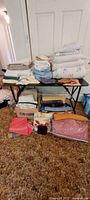 Overall view of the lot showing various folded linens including sheets, blankets, and smaller folded textile pieces placed on a table and in boxes below.