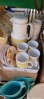 Photo showing white jug and four matching mugs with yellow stripes, small floral plates stacked.