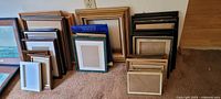 Wide assortment of photo frames of different sizes and colors, stacked and lean against wall on carpeted floor.