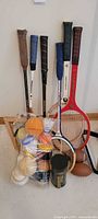 Photo shows five rackets upright against a wall with a collection of balls and shuttlecocks in packaging in front. A football is partially visible.