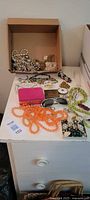 Photo of assorted vintage jewelry on a white surface including necklaces, earrings, bracelets, compacts, and sunglasses. Beaded strands in green and orange colors are visible along with pearl and floral design earrings and two small vintage compacts.