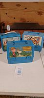 Three blue vintage kids lunch boxes featuring Snoopy & Woodstock, Lassie, and Shirt Tales characters displayed on a white table against wooden background.