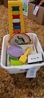 White plastic tub containing a stack of several colorful children's plastic record players, a tambourine with dancers illustration, a yellow plastic musical key-like accessory, and a vintage cassette tape.