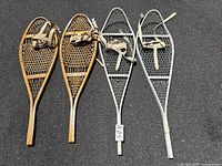 Both pairs of snowshoes laid out on pavement, showing wooden and metal frames with webbing and beige fabric straps.
