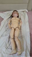 Full view of antique doll showing jointed porcelain body, nude, lying on white towel, visible paint wear and scuffs.