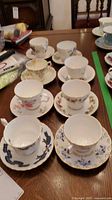 Photo showing 8 assorted teacups with matching saucers, various floral and decorative patterns, on wooden table.