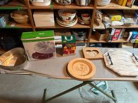Photo showing large metal baking pan, boxed kitchen gadget, wooden trivet, and some ceramic bowls on wooden shelves and a folding table.