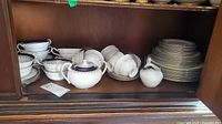 Wide view of cabinet shelf showing stacked plates, bowls, cups, saucers, sugar bowl and creamer