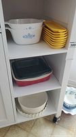 Photo of white CorningWare casserole dishes stacked with yellow rectangular Good Cook baking dishes on a shelf, along with a red and black stoneware baking dish and a white ceramic fluted tart pan below.