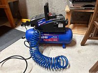 Photo showing side view of blue 3 gallon air compressor with black motor housing and attached blue coiled air hose.