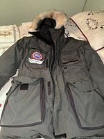 Front view of parka laid flat showing zip closure, storm flap, and multiple pockets
