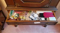 Open drawer of wooden furniture showing numerous items including taper candles, tealight candles, glass pebbles, candle holders, and miscellaneous decorative items in bags and boxes.