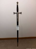 Full length view of ceremonial scabbard standing upright against a wall, showing the leather wrapped handle and metallic parts.
