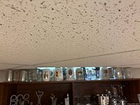 Shelf of assorted beer steins, mugs, and tankards arranged side by side under the ceiling. Mixture of glass, ceramic, and metal items with various military and commemorative designs.