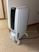 Full view of white DeLonghi oil-filled radiator heater with black control panel and caster wheels, positioned on carpet.