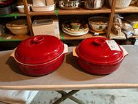 Two Emile Henry red ceramic covered casserole dishes with lids, side by side showing shape and color.