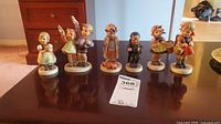 Six Goebel Hummel figurines arranged in a row on a table with a MaxSold tag visible.