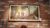 Full view of framed oil painting showing equestrian hunt scene with riders on horses and dogs in a forest landscape, hung on brick wall.